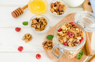 Beyaz ahşap arka planda bir cam kavanozda fındık, kurutulmuş kiraz, bal, tarçın ve yoğurt karışımı ile multicereal ev yapımı organik granola.