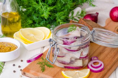 Ev yapımı salamura ringa balığı soğan, limon, taze dereotlu ve baharatlı cam bir kavanozda beyaz ahşap bir arka planda kesim tahtasında..