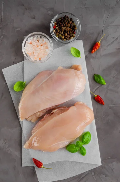 Raw chicken breast on paper with basil and other spices on a dark concrete background. Healthy food. Vertical, copy space