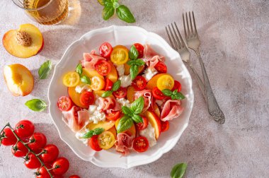 Mozzarella soslu salata, şeftali, jambon, domates ve beton arka planda beyaz bir tabakta taze fesleğen. Üst görünüm, boşluğu kopyala. Sert ışık