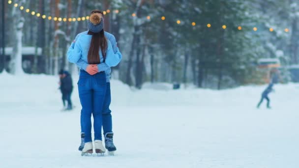 Αγκαλιές ζευγάρι στέκεται σε υπαίθριο παγοδρόμιο με γιρλάντες — Αρχείο Βίντεο