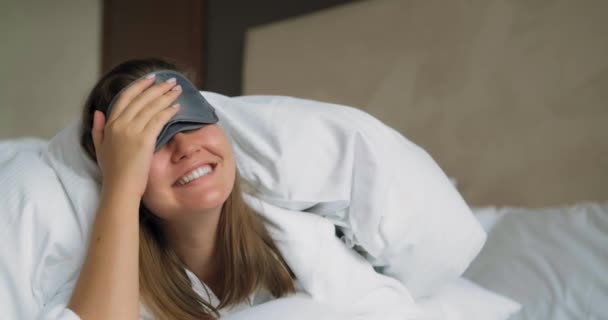 침대 위의 부드러운 담요 밑에 누워 있는 여성의 수면 마스크를 벗는다 — 비디오