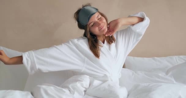 Красива жінка розтягується і одягає маску для сну, сидячи на ліжку — стокове відео