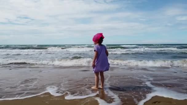 Child on ad the sea beach with rough sea have fun walking and jumping with a bandana and purple dress — Stock Video