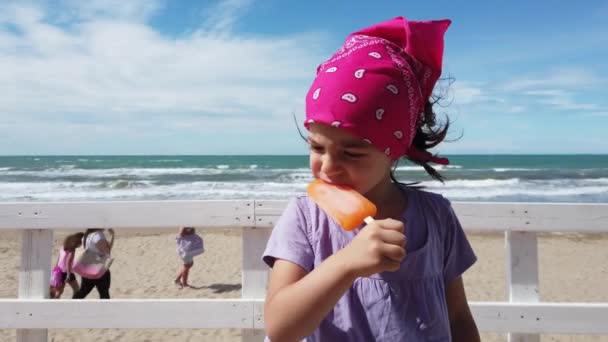 Dziewczynka dziecko gryzienie lodów lodów na plaży bar wietrzny morze różowy bandana — Wideo stockowe