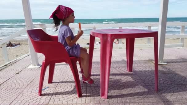 Linda niña comiendo popcicle en el bar de la playa en la silla roja y mesa — Vídeos de Stock