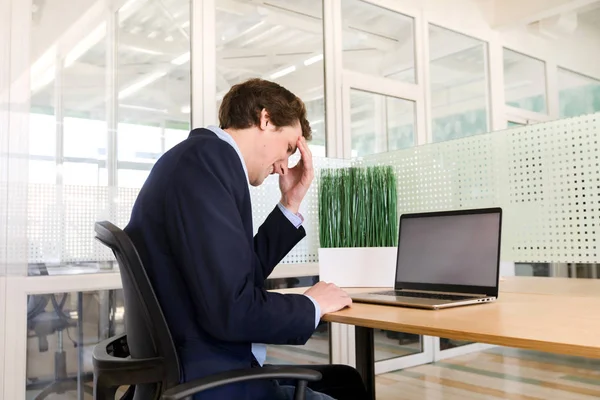 Young man in suit sitting at table in office and brainstorming deeply having laptop on top in front