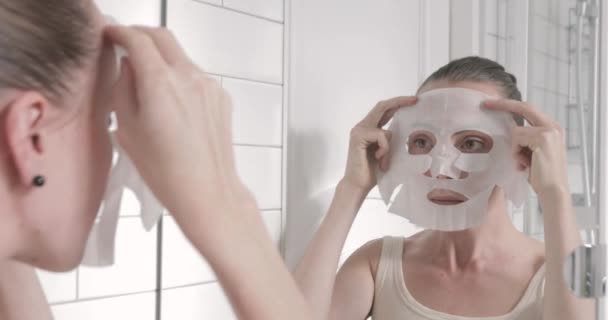 Женщина надевает маску на лицо, глядя в зеркало — стоковое видео