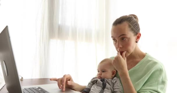テーブルで赤ちゃんと働く母親 赤ちゃんを手にノートパソコンで働く忙しい女性 居心地の良い家で手に美しい幼児と働くお母さん 女性のフリーランスの仕事 現代の母親 — ストック動画