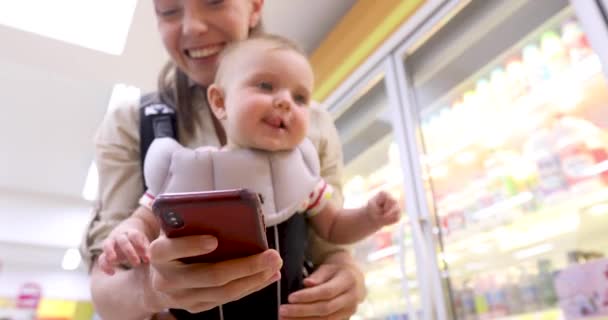 Mujer con bebé en cabestrillo utiliza el teléfono inteligente en la tienda — Vídeo de stock