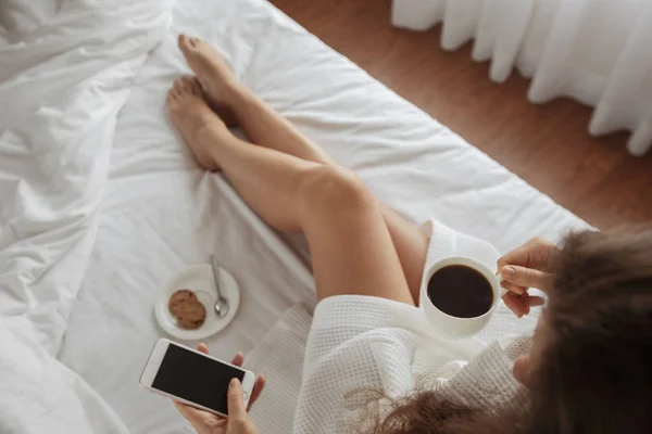 Flicka ligger säng dricker hennes morgon kaffe ser telefon — Stockfoto