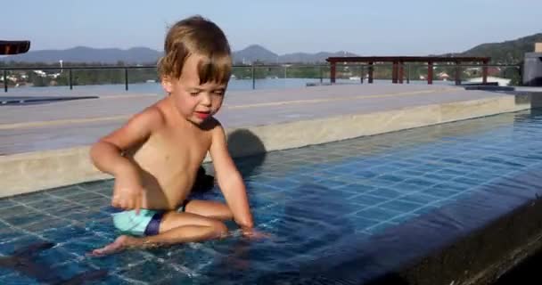 Mały chłopiec siedzi na brzegu i rzuca kamieniami w wodę na zewnątrz — Wideo stockowe