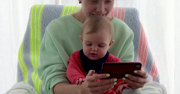 Madre con asientos de hija pequeña cerca de la ventana y utiliza el teléfono inteligente — Vídeos de Stock