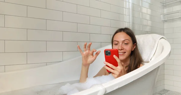 Брюнетка в ванной со смартфоном в пене — стоковое фото