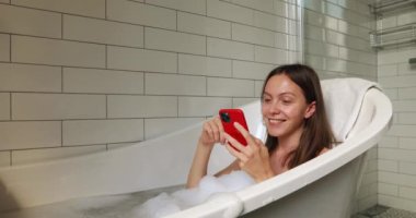 Küvette köpüklü akıllı telefonlu esmer bir kadın.