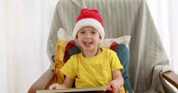 Chico divertido en rojo blanco sombrero de Navidad se sienta en la silla sonriendo — Vídeos de Stock