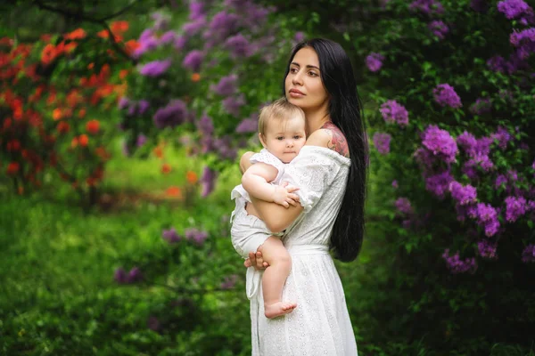 Uma jovem mãe segura um bebê em seus braços contra o fundo de um arbusto florido — Fotografia de Stock