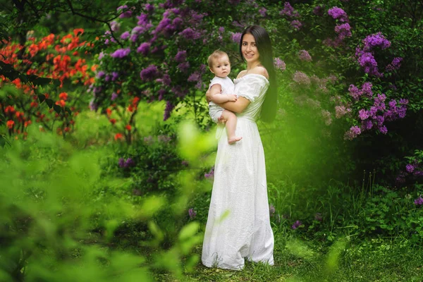 A jovem mãe segura sua filhinha em seus braços entre árvores florescentes. Árvore lilás. Dia das mães — Fotografia de Stock