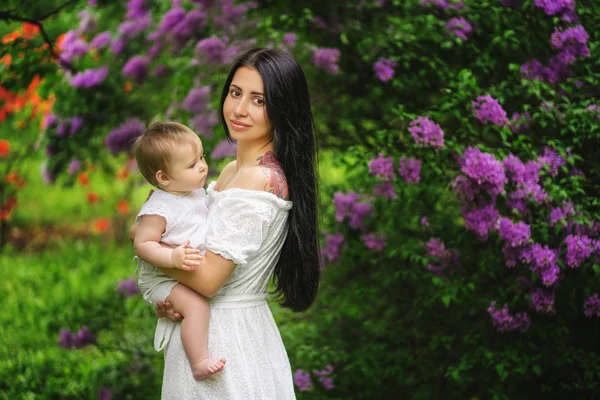 Giovane madre tiene la sua figlioletta tra le braccia tra gli alberi in fiore. Albero lilla sullo sfondo — Foto Stock