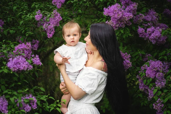 Menina bebê em mãos mãe olha para a moldura. Árvores florescentes ao redor — Fotografia de Stock