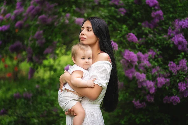 Mamma tiene il bambino tra le braccia. Il bambino guarda nella cornice. Donna che distoglie lo sguardo. Albero in fiore — Foto Stock