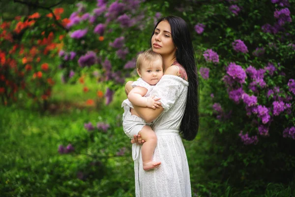 Mom and her baby in white clothes on the background of flowering trees and bushes. — Stock Photo, Image