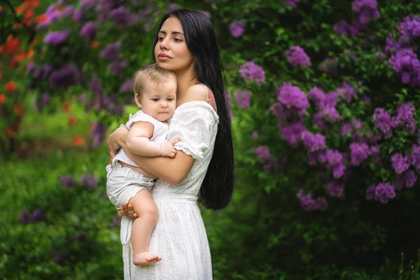 Woman in a white dress with her little daughter on the hands .Background of flowering trees. — Stock Photo, Image