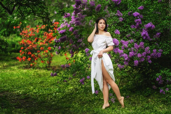 Barefoot dziewczyna w białej sukni spacery w ukwieconym ogrodzie. Krzewy Lilac — Zdjęcie stockowe