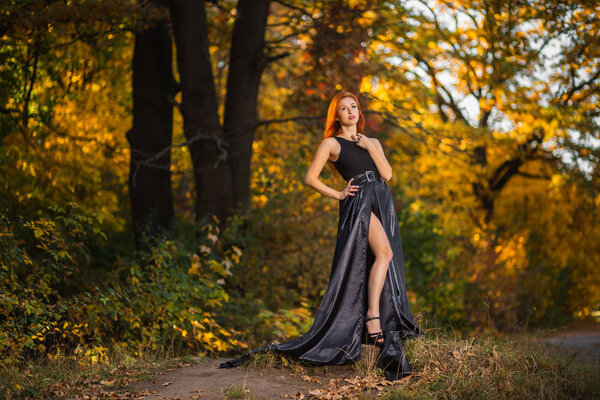 Beautiful woman with long red hair posing in a black dress with a long train standing in a autumn forest.