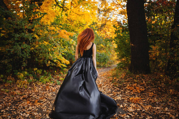 Cute girl with long red hair wearing a black dress spinning and running in autumn orange forest. no face.