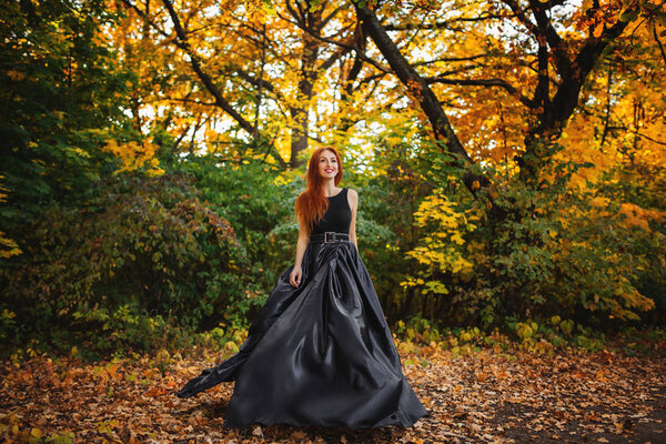 Amazing red haired model in black dress looking at camera and posing. Autumn nature at background