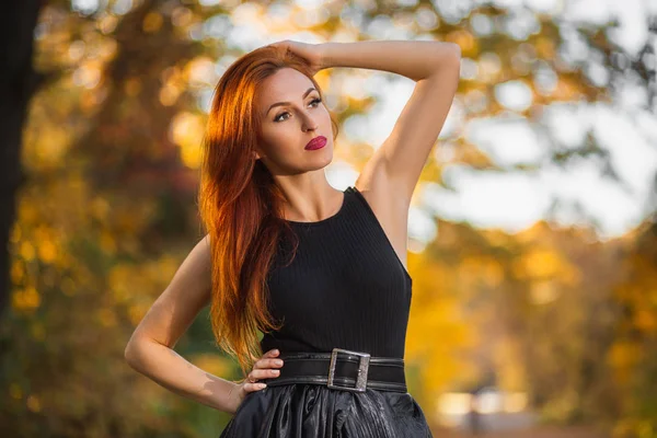 Close up portrait of a beautiful redhead girl in dark dress standing near colorful autumn leaves. Stockfoto