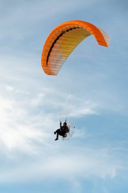 Mavi gökyüzüne karşı paramotor uçuşu, sarı ve turuncu bir gölgeliğin altında asılı bir adamın silüeti.