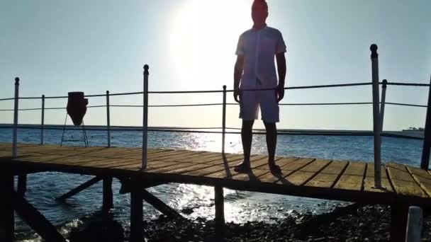 El hombre levanta las manos para saludar. Un hombre se para en un muelle cerca del mar — Vídeos de Stock