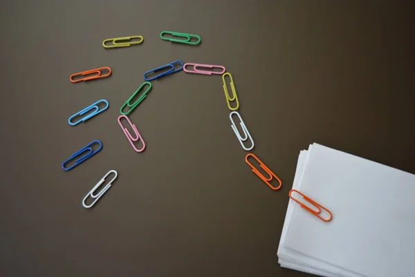 Colored paper clips laid out with a question mark and with sheets of white paper for recording on a brown matte background.