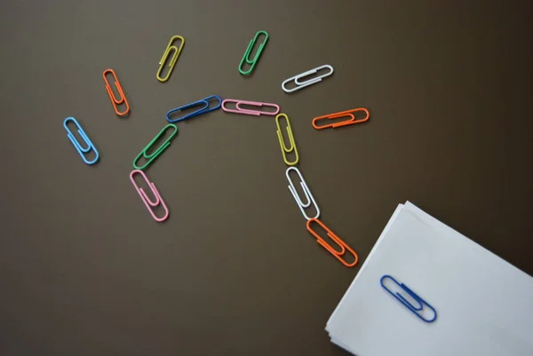 Colored paper clips laid out with a question mark and with sheets of white paper for recording on a brown matte background.