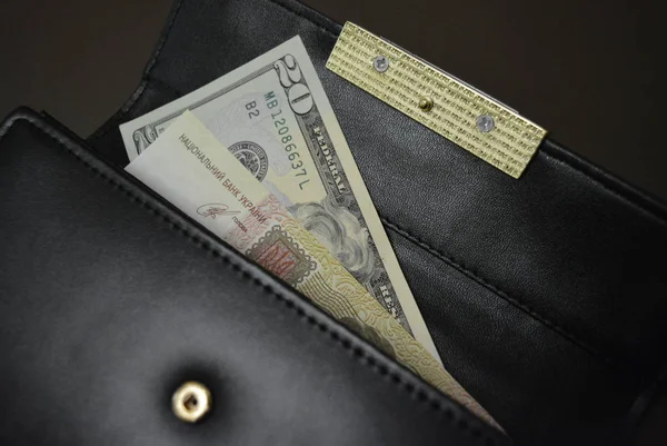 Black leather wallet with money in it on a brown matte background. Banknotes 20 twenty American dollars and 100 one hundred Ukrainian hryvnias inside a black wallet.wallet,