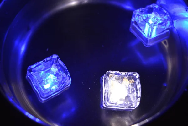 Plastic glowing bright blue & white ice cubes in water. Led ice floes are floating in a stainless steel dipper with an interesting metal reflection.