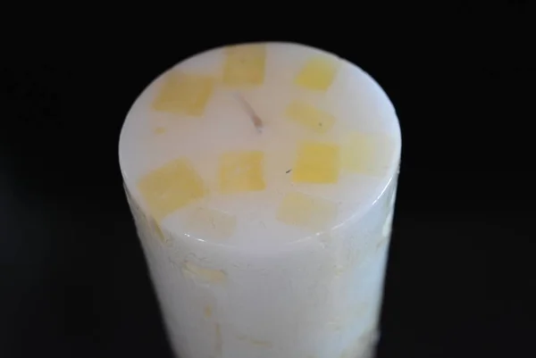 White, with yellow squares, a round, long, thick candle with a vanilla aroma is located on a black glossy surface.