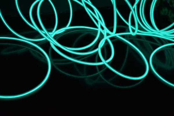 Unusual bright wires made of turquoise-colored material glowing at night. Chaotic sky blue wires, light guide electroluminescent wires, electroluminescence are located on a glossy black surface.