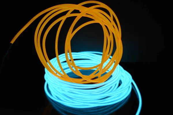 A bright coil of turquoise, sky blue luminous wire with chaotic wires of an orange light guide located on a black glossy surface. Light canvas, backlighting with wires and art background.