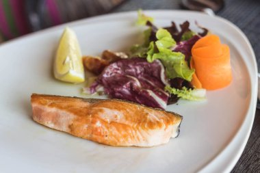 Beyaz tabakta ızgara somon, sebze ve limonla servis edilir..