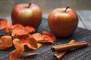 Olgun kırmızı elmalar ve turuncu sonbahar yaprakları tarçın çubuklarının yanında koyu bir zemin üzerinde.