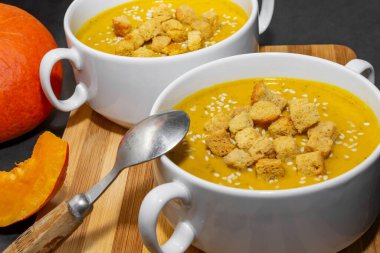 Ahşap zemin üzerinde susam tohumları olan sonbahar balkabağı çorbası. İki kase portakal çorbası ve arka planda balkabağı dilimleri.