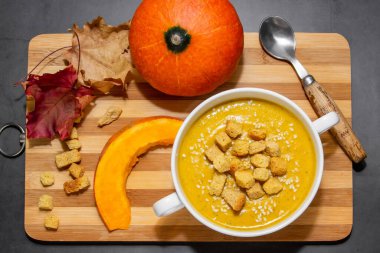 Ahşap zemin üzerinde susam tohumları olan sonbahar balkabağı çorbası. Portakal rengi sonbahar balkabağı çorbası ve kaşık arka planda ağaç ve yapraklar.