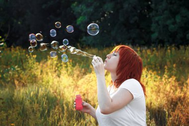 Genç kızıl saçlı kadın sabun köpüğü üflüyor. Doğada mutlu bir kız.