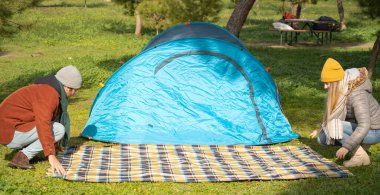 Bir çift doğada kamp alanında kamp çadırı ve piknik battaniyesi kuruyor.