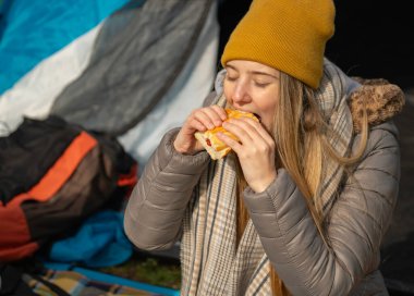 Kamp gezisi sırasında çadırın önünde İspanyol sucuklu sandviç yiyen kampçı kızın portresi.
