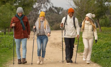 Dört mutlu yürüyüşçüden oluşan çok ırklı bir grup, yürüyüş sırıkları kullanarak bir patikada yürüyor ve konuşuyorlar.