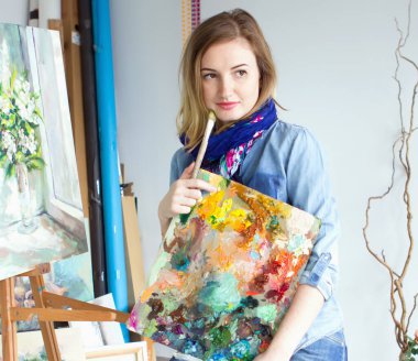 Elinde boya paleti olan bir kız sanatçı. Doğrudan kameraya bakıyor. Sanat stüdyosu